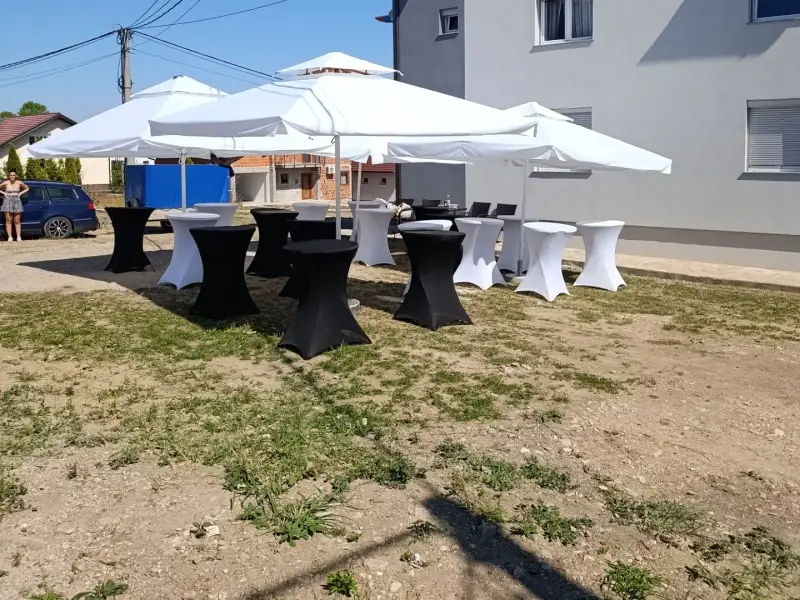 Barski stolovi s bijelim i crnim navlakama ispod bijelih suncobrana ispred zgrade